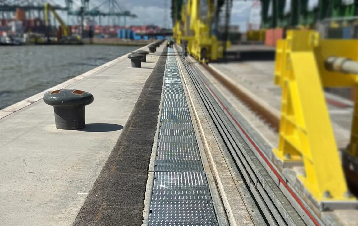  Blick entlang einer Hafenpier mit Pollern am Wasser; mittig die Abdeckung des Grabens, in dem die iMSPO verfährt, rechts gelbe Krananlagen und Schienen, im Hintergrund Container und Kräne.