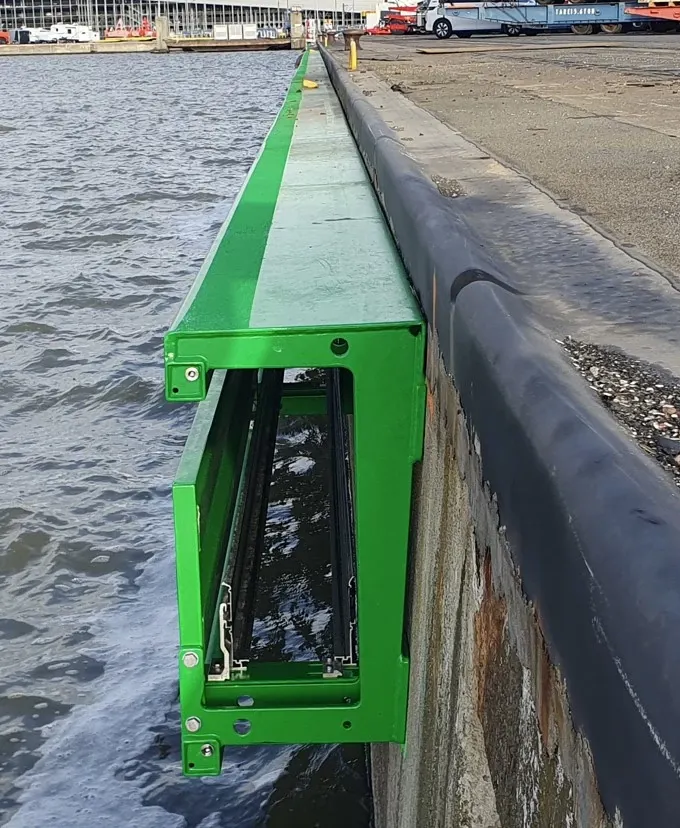 Auf dem Bild ist eine Kaimauer zu sehen. Im Hintergrund sieht man ein Containerschiff am Kai, das gerade von mehreren Kranen entladen wird. An der Kaimauer entlang ist eine gelbe Struktur montiert. Hierbei handet es sich um eine an der Kaimauer entlang montierte Landstromlösung - die iMSPO QF. 